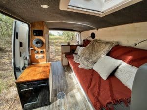 L'image dépeint l'intérieur chaleureux et invitant d'un van aménagé, conçu pour le voyage et l'habitation. L'espace est optimisé avec un grand lit confortable recouvert d'une couverture rouge et de coussins blancs, créant une atmosphère accueillante. À gauche, un meuble en bois fait office de cuisine compacte, équipée d'un réfrigérateur noir moderne et d'un plan de travail brillant. Au-dessus, une série de haut-parleurs intégrés promet une ambiance musicale pour les routes. Des touches de finition en bois sur les portes et les placards ajoutent une sensation de confort domestique. La vue par la porte arrière ouverte montre un paysage naturel extérieur, invitant à l'exploration et à l'aventure en plein air