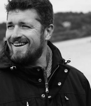 Photographie en noir et blanc d'un homme affichant un sourire sincère et joyeux. Il porte une veste d'extérieur noire avec des détails de cordon, adaptée pour une ambiance maritime ou hivernale. Le sujet, qui pourrait être identifié comme Gilles selon le contexte donné, semble regarder quelque chose ou quelqu'un hors cadre, ce qui ajoute une touche de spontanéité à l'image. Le flou doux de l'arrière-plan met en exergue son visage expressif, capturant un moment de bonheur ou de réaction agréable. Cette image dépeint un caractère affable et un esprit aventureux.