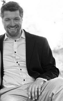 Portrait en noir et blanc d'un homme nommé Gilles, capturé avec un sourire chaleureux et accueillant. Il est assis, vêtu élégamment d'un costume noir avec une chemise blanche, la veste ouverte pour une touche de décontraction. L'image évoque une atmosphère de professionnalisme détendu. Gilles est présenté comme le fondateur de Letivan, suggérant qu'il est un entrepreneur décontracté mais sérieux. La lumière naturelle derrière lui crée un effet de halo, mettant en valeur son expression amicale et accessible